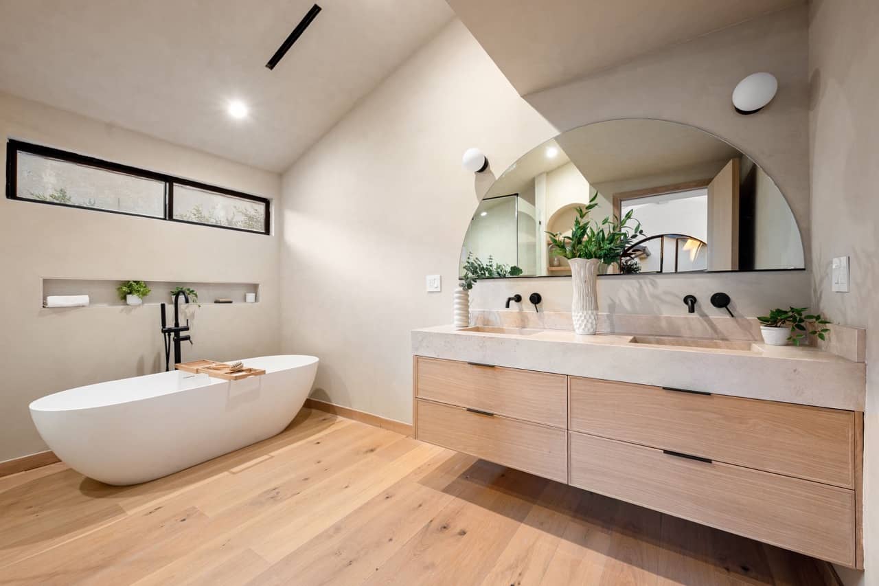 Freestanding tub under clerestory windows beside a wood vanity with an arched mirror