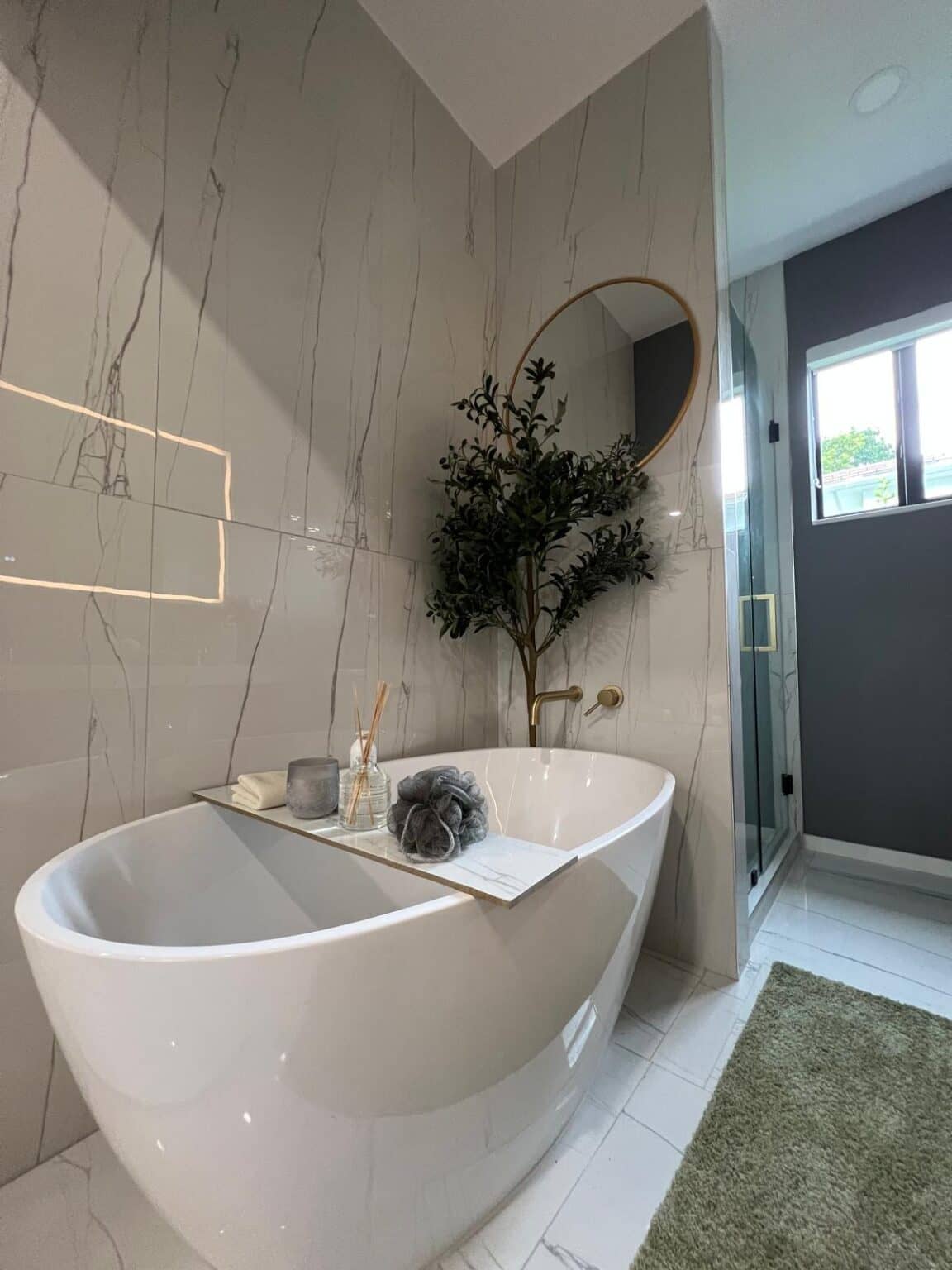 Freestanding tub against a marble-look porcelain wall with a lit niche and a round mirror